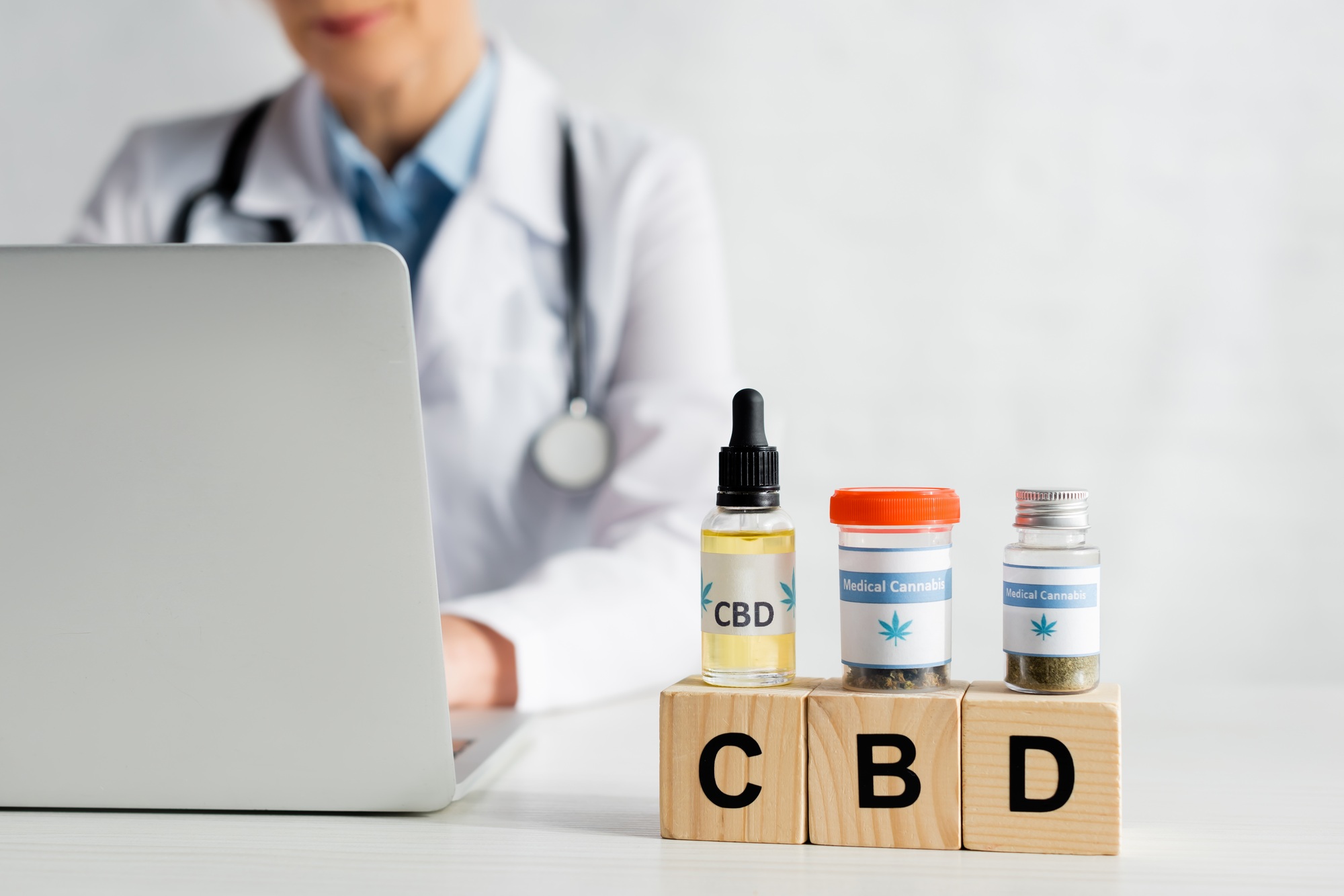 cropped view of doctor using laptop near cubes with cbd lettering and bottles with medical cannabis