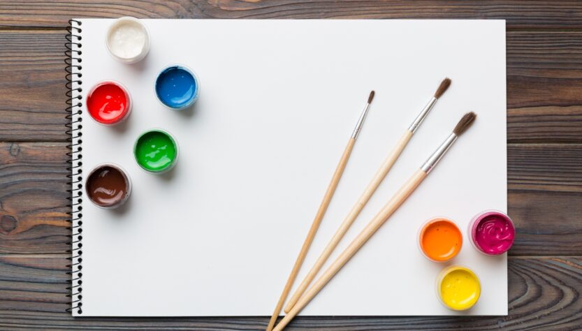 Album pages and paints, pencils, brush on a colored background. Top view with empty space